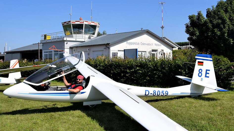 Pilot im Cockpit eines Segelflugzeugs vor einem Flugplatzgebäude